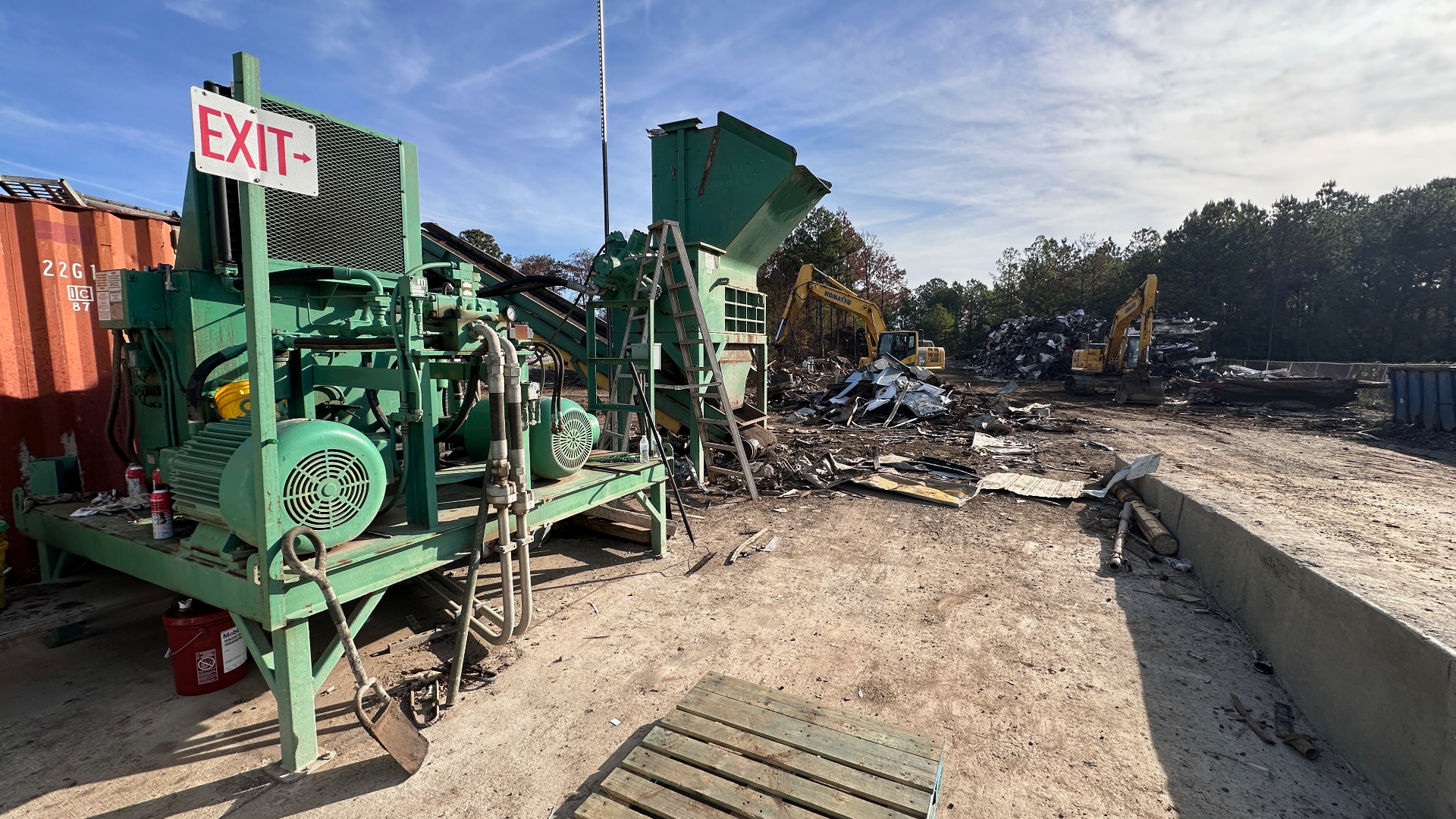 Wide shot of the CW Mobile Service processing yard: green two-ram baler and shear-shredder with conveyor, feeding pile of prepared ferrous scrap while Komatsu excavators work the pile in the background